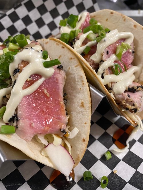 Three tacos filled with seared tuna, drizzled with sauce, topped with green onions and served in a metal holder.