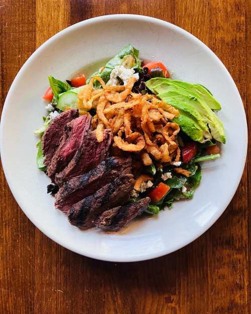 A plate of salad topped with grilled steak, avocado slices, tomatoes, and crispy onion rings on a wooden table.