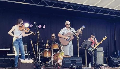 A band performs on stage, featuring a singer with an acoustic guitar, a violinist, and a drummer.