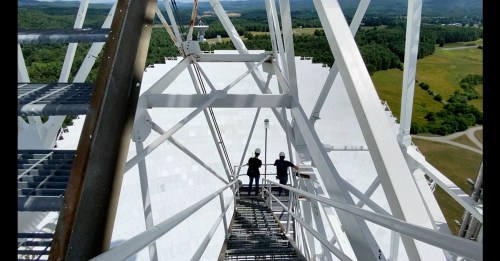Two people standing on a metal staircase of a large structure, overlooking a green landscape below.