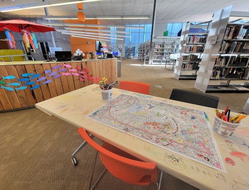 A colorful library space featuring a decorated table with art supplies and a large coloring page, surrounded by bookshelves.