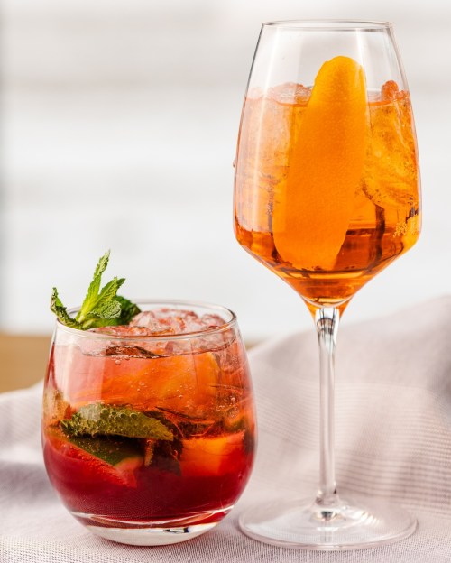 Two colorful cocktails: one in a short glass with mint and fruit, the other in a tall glass with an orange slice.