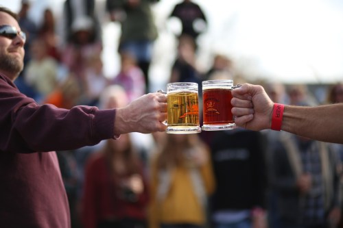 Two people clink beer mugs together in a lively outdoor setting, with a crowd blurred in the background.