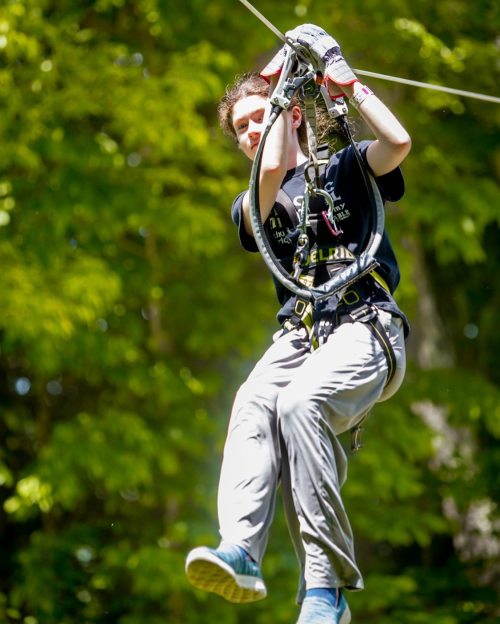 A person in a harness zip-lining through a forest, wearing gloves and casual clothing, with greenery in the background.