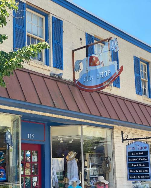 A storefront of "The Fun Shop" featuring a rocking horse sign, blue shutters, and a display of children's items.