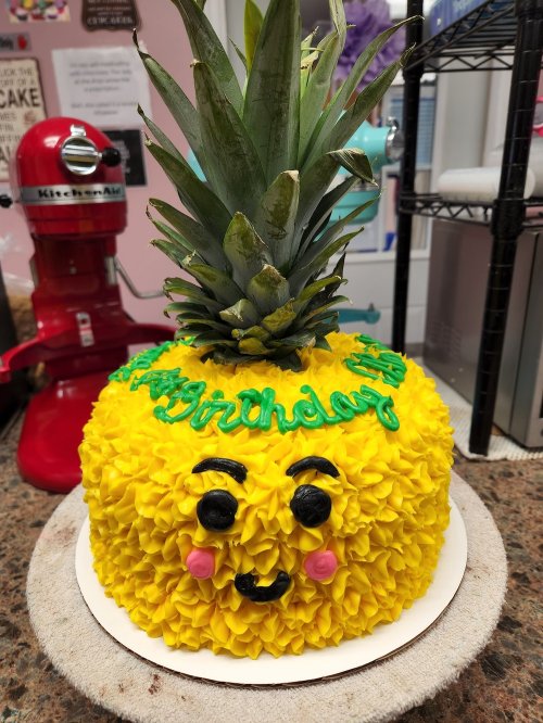 A cake shaped like a pineapple, decorated with yellow frosting, a green top, and a smiling face.