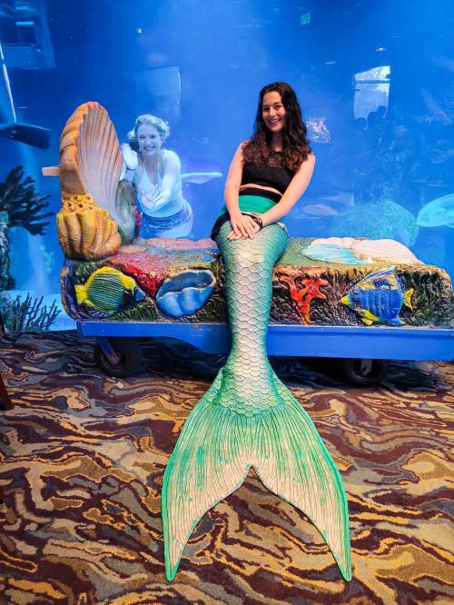 A woman in a mermaid costume poses on a colorful sea-themed bench, with an aquarium backdrop and another woman behind her.