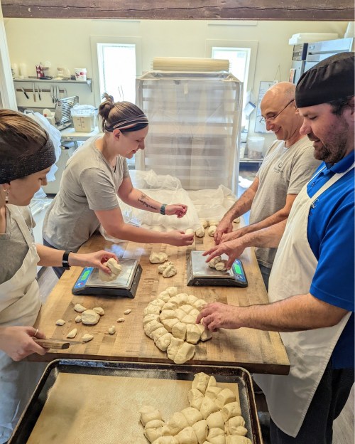 Four bakers work together in a kitchen, shaping and weighing dough on a wooden table. Flour and baking tools are visible.