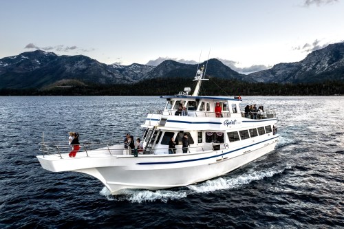 A white boat with blue accents sails on a lake, surrounded by mountains and a clear sky, with people on board enjoying the view.