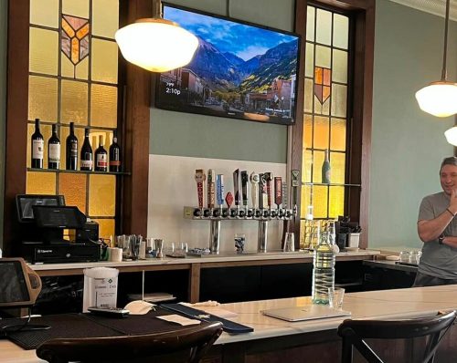 A modern bar with patrons, a TV on the wall, and stained glass windows, featuring a sleek counter and seating.
