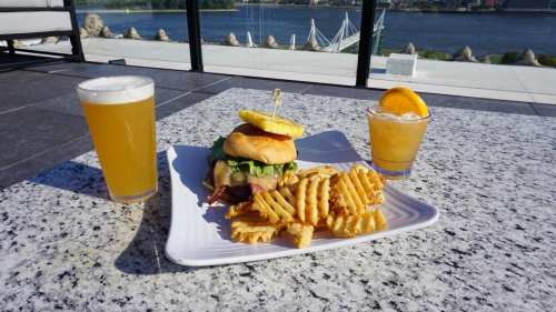 A burger with lettuce and pineapple, served with waffle fries, alongside two drinks, with a scenic view in the background.