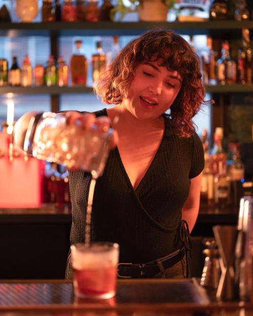 A bartender with curly hair pours a drink into a glass at a well-stocked bar, focused and engaged in her work.