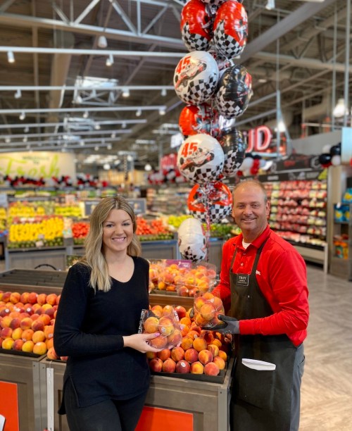 A woman and a man hold peaches in a grocery store, surrounded by colorful fruit displays and festive balloons.