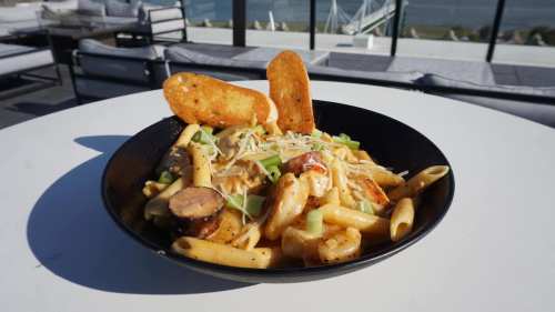 A bowl of creamy pasta with shrimp, sausage, and green onions, garnished with crispy breadsticks, set against a scenic backdrop.