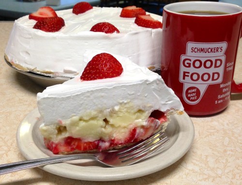 A slice of strawberry cream pie on a plate next to a red mug of coffee, with the whole pie in the background.