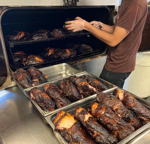 A person wearing gloves removes smoked meat from a large smoker, with trays of cooked meat in the foreground.