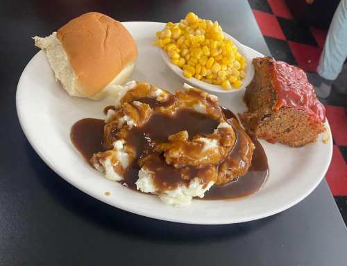 A plate with meatloaf topped with sauce, mashed potatoes with gravy, corn, and a dinner roll.