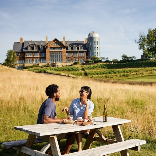 A couple enjoys a picnic at a scenic outdoor table with a large building in the background.