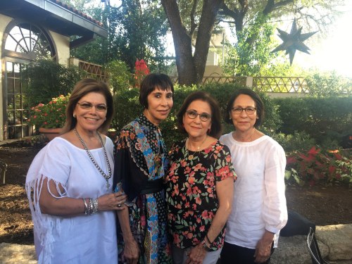 Four women pose together outdoors, surrounded by greenery and decorative elements, smiling at the camera.