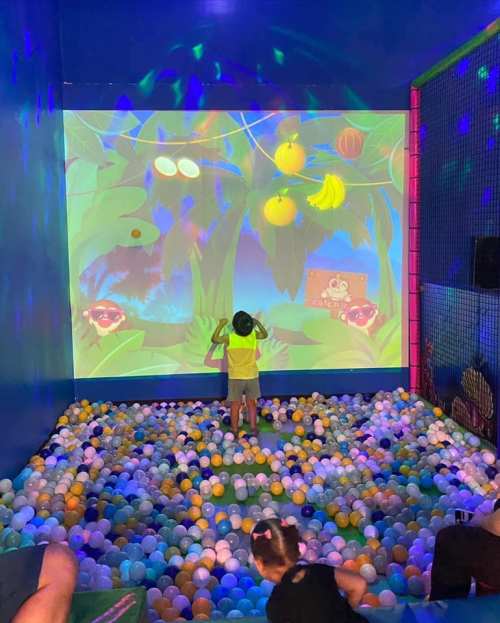 A child plays in a colorful ball pit, interacting with a vibrant projection of a jungle scene on the wall.