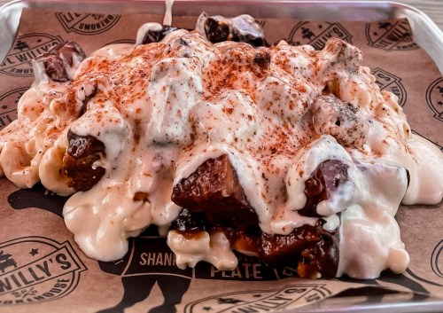 A close-up of a plate of mac and cheese topped with smoky meat and drizzled with creamy sauce and spices.
