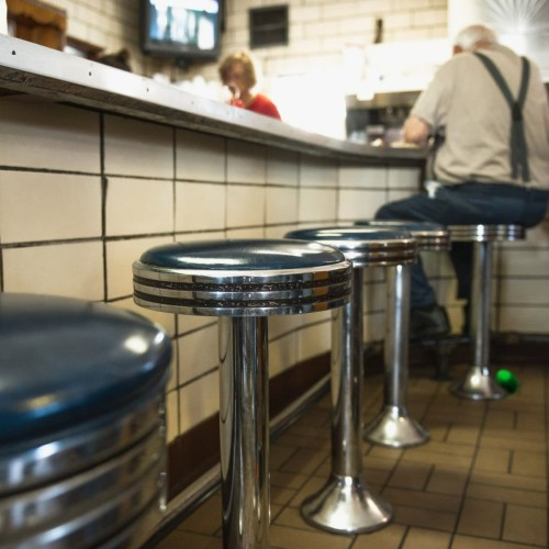A retro diner scene featuring chrome stools and a tiled counter, with a person sitting in the background.