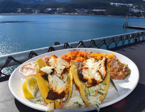 A plate of fish tacos with slaw, rice, and beans, set against a scenic waterfront view.
