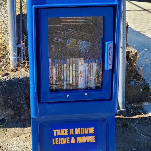 A blue movie exchange box with a glass front displaying DVDs, labeled "Take a Movie, Leave a Movie."