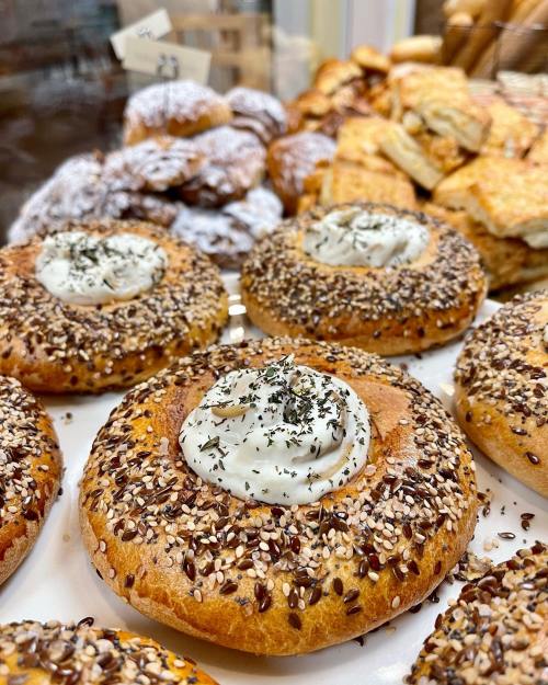 A close-up of bagels topped with cream cheese and sprinkled with seeds, surrounded by various baked goods.