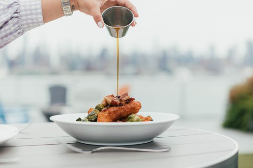 A hand pours sauce over a plate of fried food and vegetables, with a city skyline in the background.