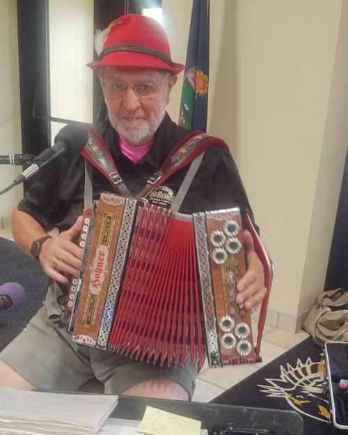 A man in a red hat and shirt plays an accordion while sitting at a table with a microphone.