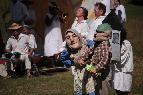 A group of performers in white costumes, featuring large puppets, play music outdoors in a sunny setting.