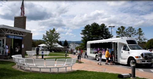A tour center with visitors boarding a shuttle bus on a sunny day, surrounded by trees and mountains in the background.