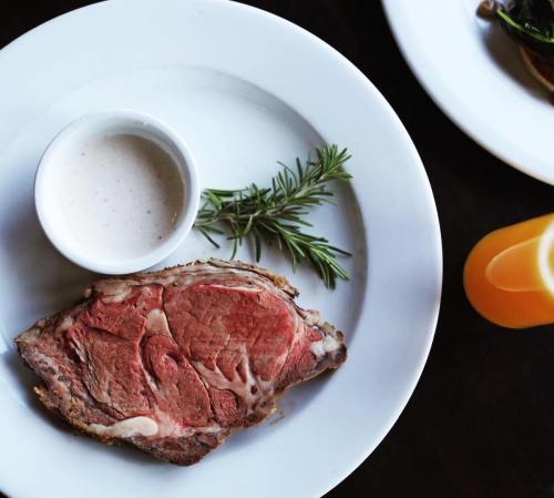 A slice of roast beef on a plate with a small bowl of sauce and a sprig of rosemary.
