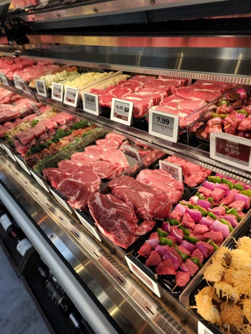 A display of various meats in a grocery store, including steaks, chops, and marinated options, with price tags visible.
