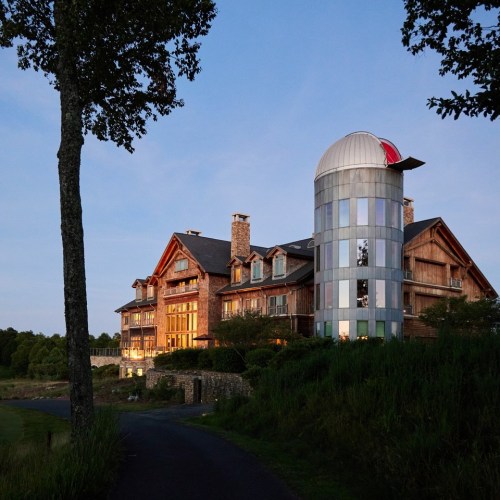 A modern building with a glass tower next to a rustic lodge, surrounded by greenery and a winding path.