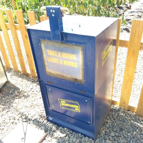 A blue movie exchange box with "Take a Movie, Leave a Movie" text, surrounded by a wooden fence and gravel.