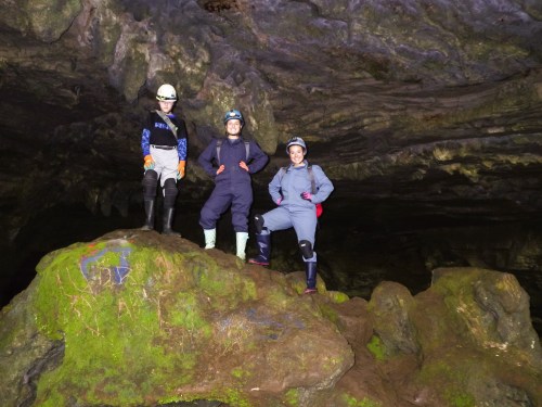 Three people in helmets and protective gear pose on a mossy rock inside a cave.