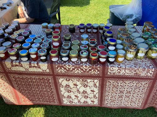 A table covered with a patterned cloth displaying various jars of colorful jams and jellies at an outdoor market.