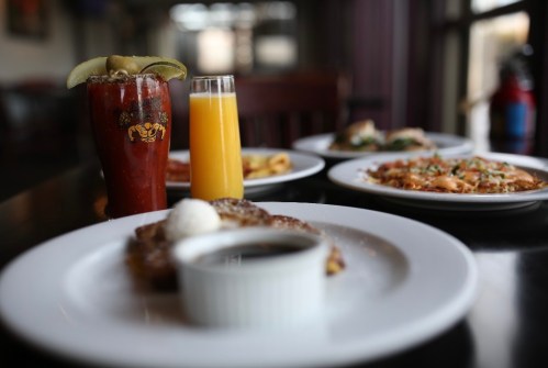 A table set with a Bloody Mary, mimosa, and various breakfast dishes, including pancakes and eggs, in a restaurant setting.