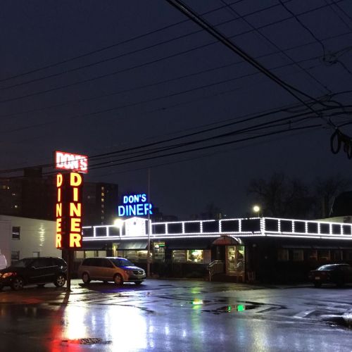A diner at night, illuminated with neon signs and festive lights, reflecting on wet pavement.