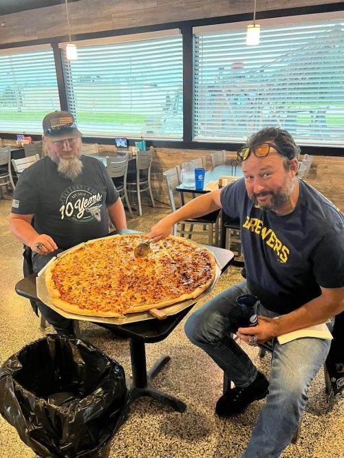 Two men sitting at a table with a large pizza, one slicing it while the other watches, in a casual dining setting.