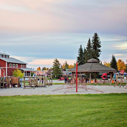 A playground with swings and climbing structures, surrounded by trees and buildings under a colorful sky at sunset.