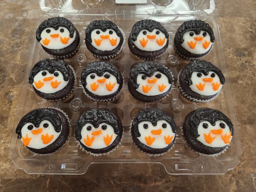 A dozen cupcakes decorated as penguins with black frosting, white icing, and orange beaks, arranged in a clear container.