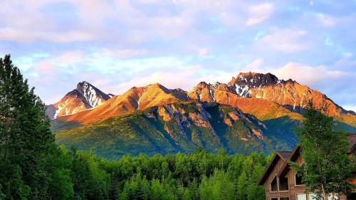 Sunlit mountains with snow-capped peaks rise above a lush green forest and a wooden building in the foreground.