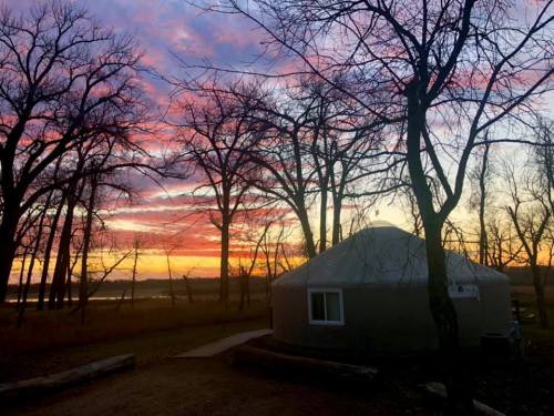 A yurt surrounded by bare trees at sunset, with vibrant pink and orange clouds in the sky.