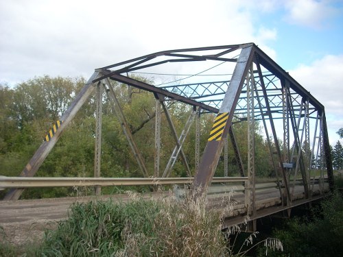 A metal truss bridge spans a river, surrounded by trees and under a cloudy sky.