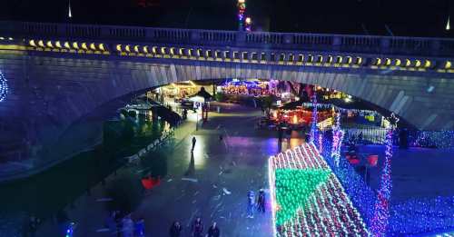 A vibrant night scene of a bridge adorned with colorful lights, overlooking a festive market below.