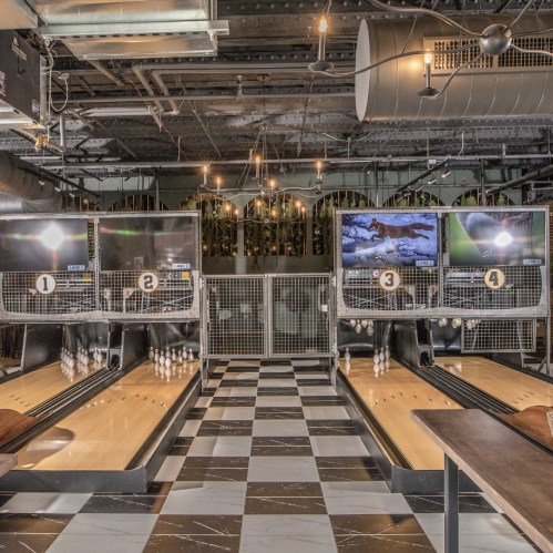A modern bowling alley with four lanes, screens above, and stylish decor featuring checkered flooring and industrial lighting.