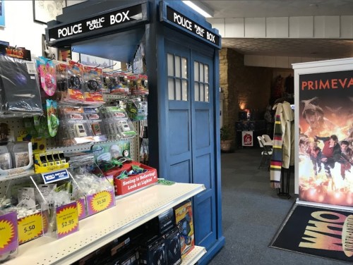 A blue police box stands in a store, surrounded by toys and merchandise, with a promotional display in the background.